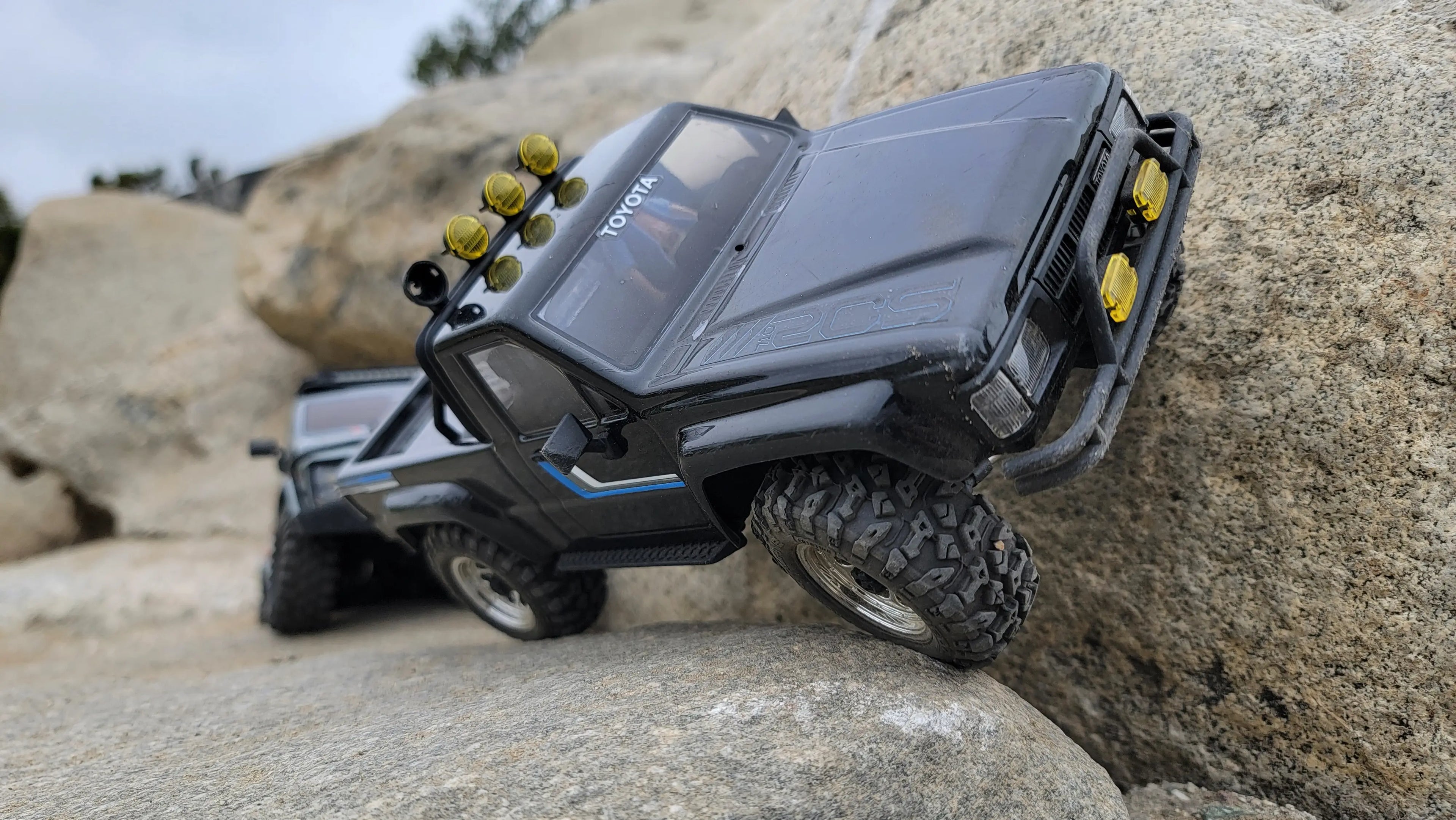 Black Toyota RC crawler with yellow lights scaling boulders, featuring RCSTEVE performance parts for realism and trail performance.