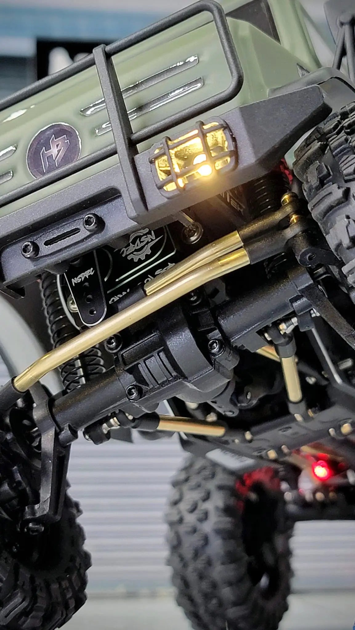 Close-up of RCSTEVE brass link upgrade on an RC crawler, showing detailed suspension components, NSDRC servo, and LED lighting for high-performance crawling.